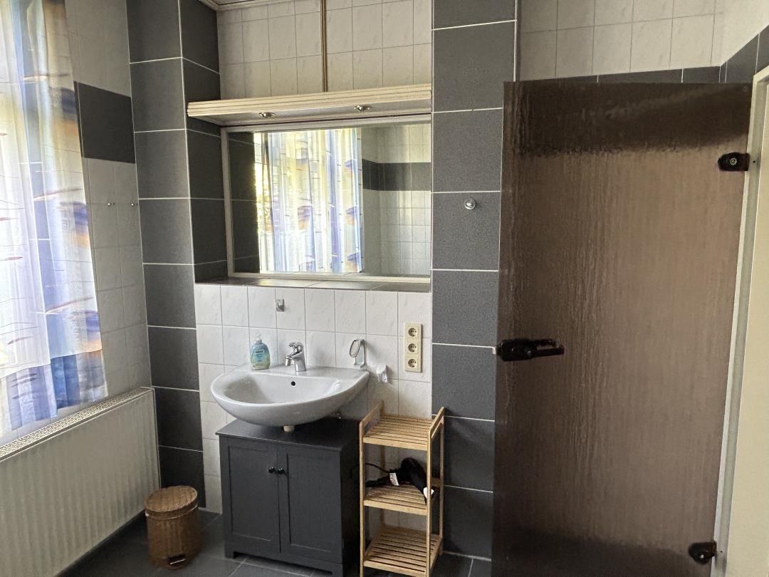 Badezimmer mit Waschbecken, grauem Unterschrank, Spiegel und Holzregal.