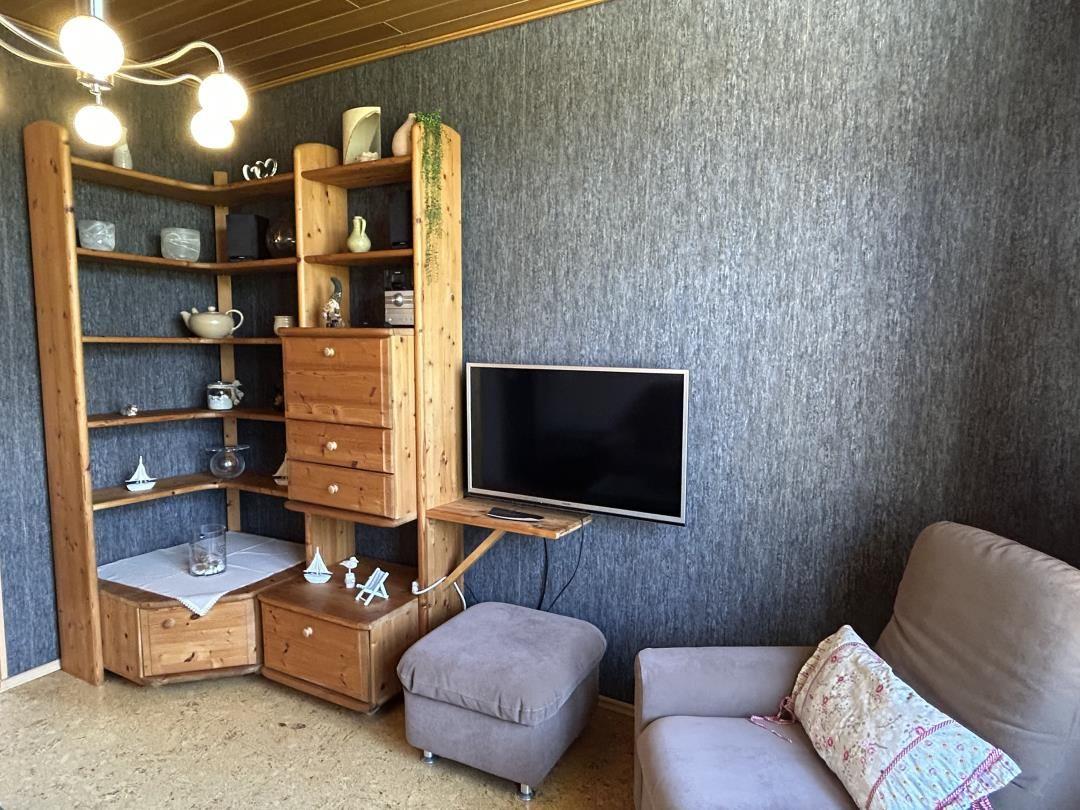 Wohnecke mit Holzregal, Wand-TV, grauem Sessel und Hocker.