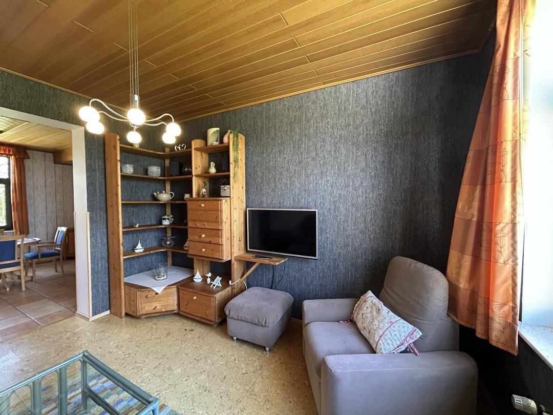 Wohnbereich mit grauem Sessel, TV und Holzschrankwand an dunkler Wand.