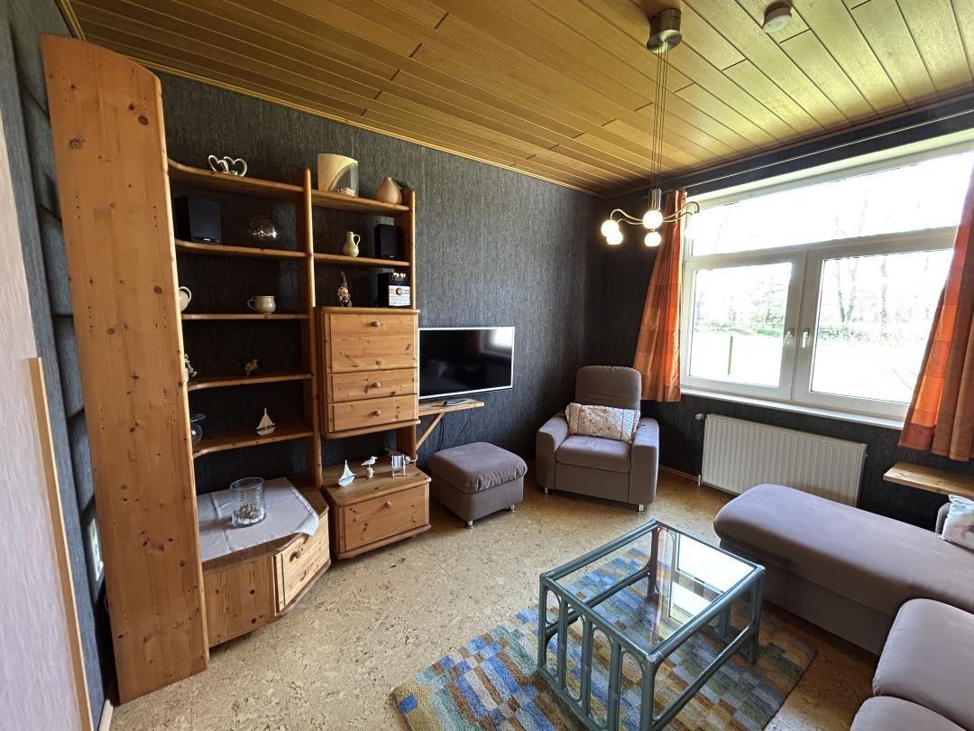 Wohnzimmer mit Holzdecke, Schrankwand, Sessel, Glastisch und TV.
