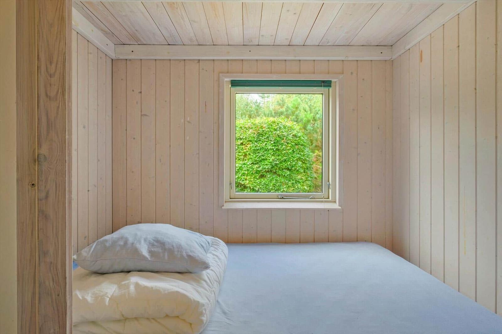 Kleines Schlafzimmer mit Holzwänden, Bett und quadratischem Fenster ins Grüne.