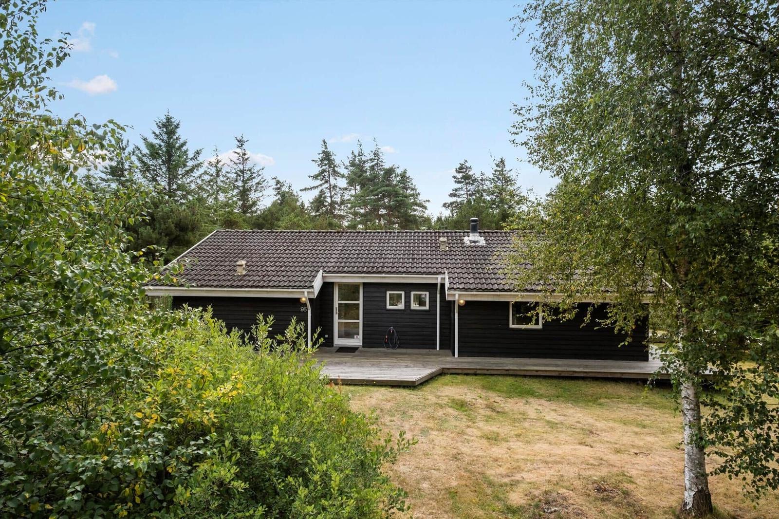 Dunkles Holzhaus mit Satteldach, weißer Tür und Holzterrasse vor Bäumen.