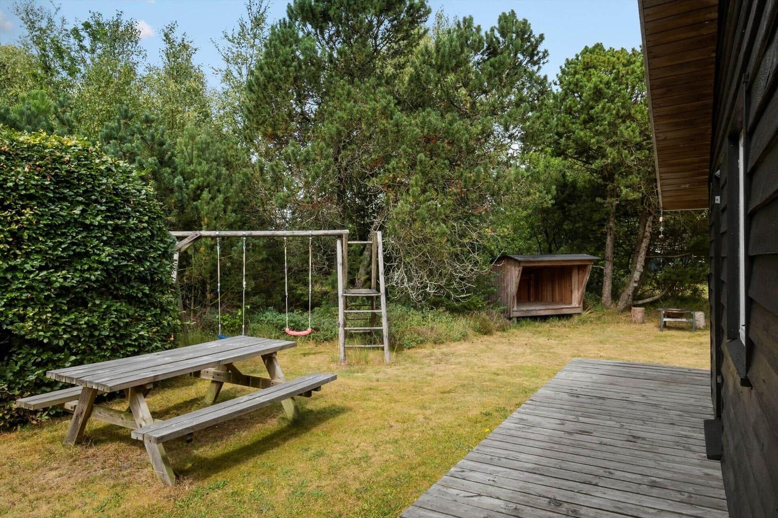 Garten mit Holzterrasse, Picknicktisch, Schaukel und Holzunterstand vor Bäumen.