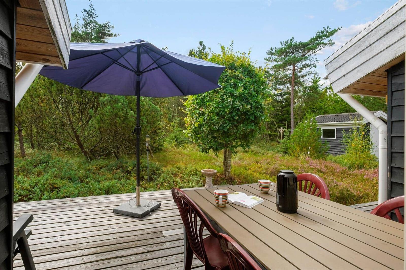 Holzterrasse mit Esstisch, roten Stühlen, blauem Schirm und Gartenhaus.