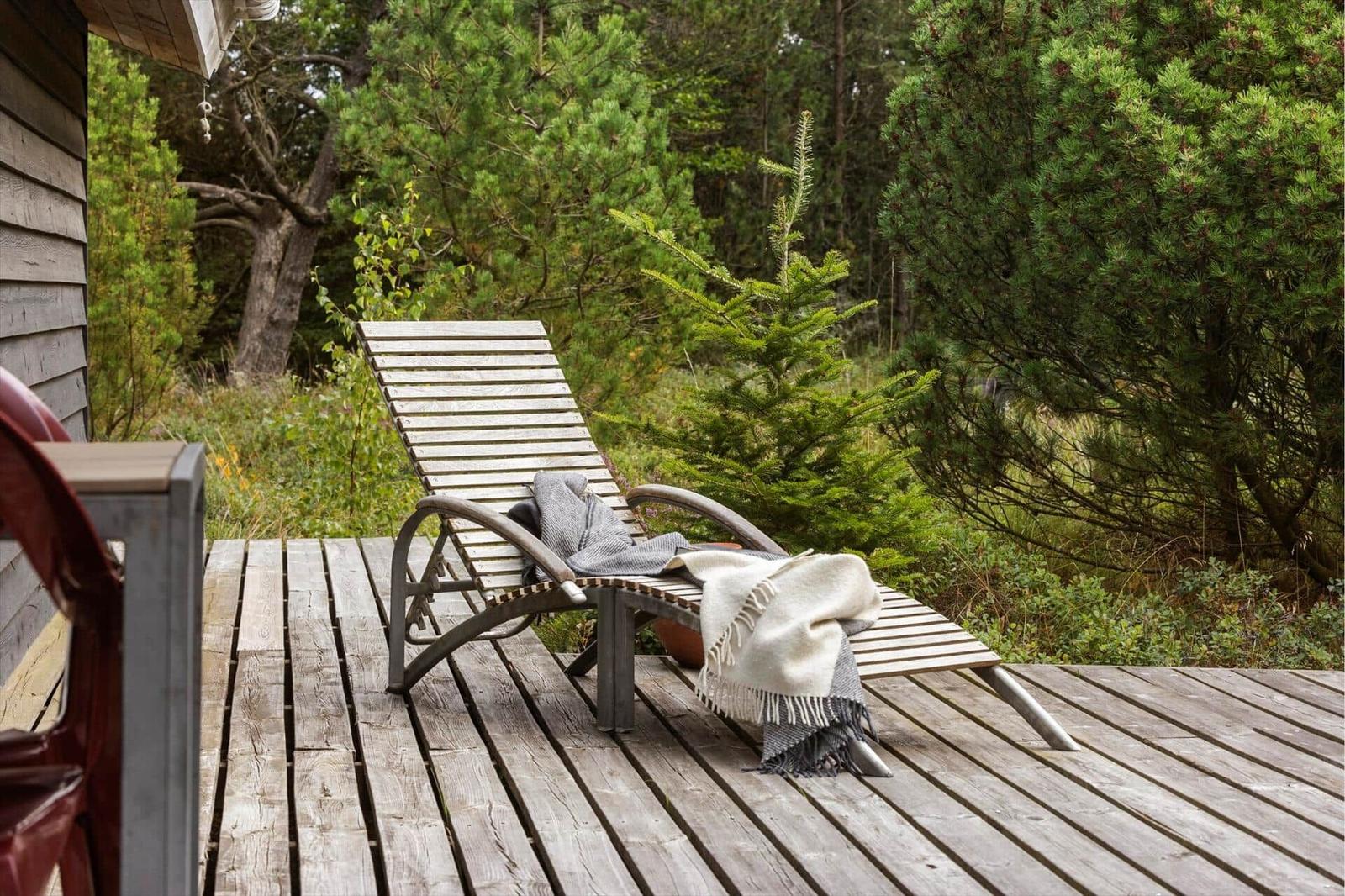 Holzliege mit Decke auf Holzterrasse vor Tannen und Sträuchern.
