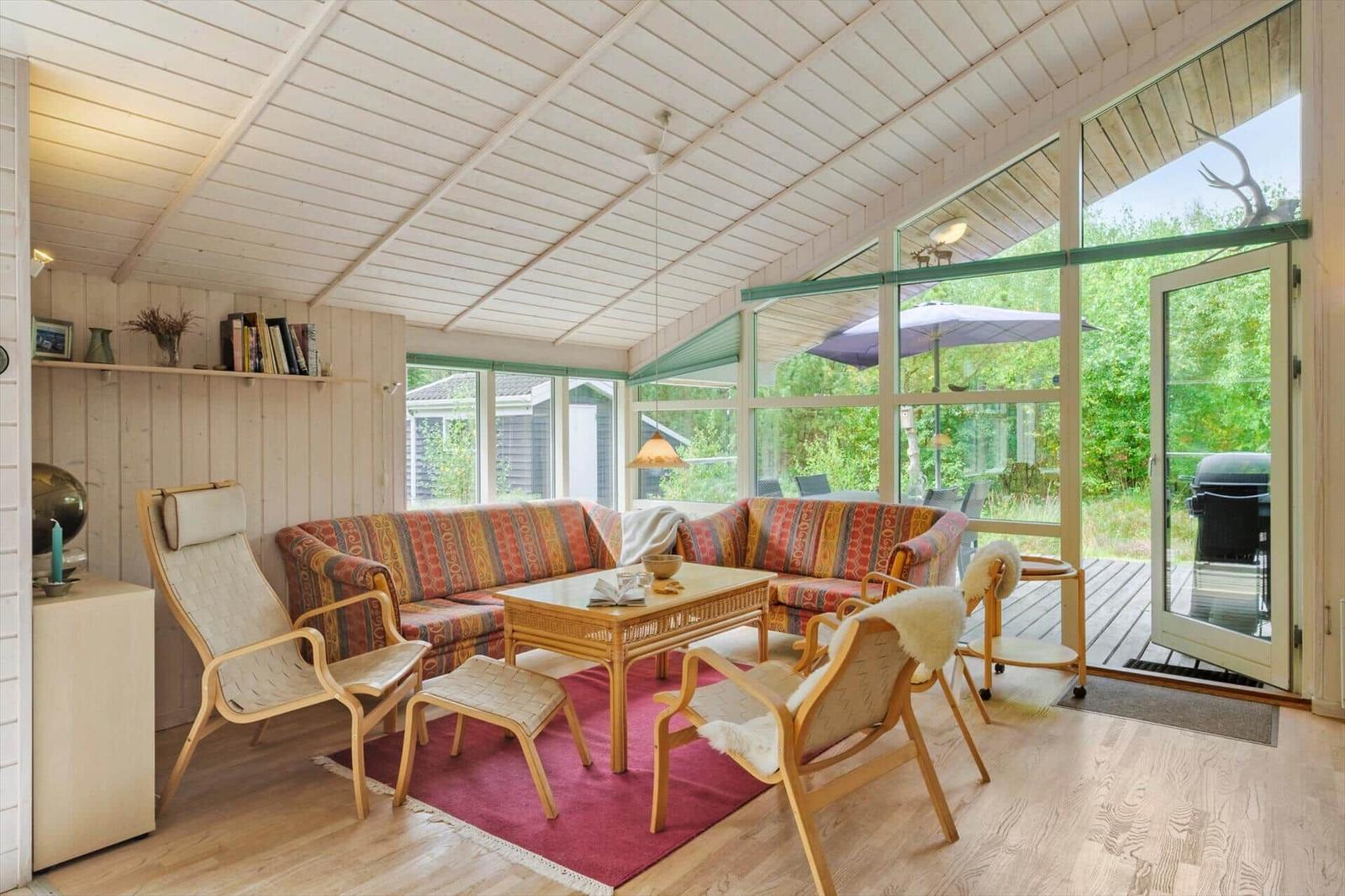 Wohnzimmer mit Ecksofa, Sesseln, Couchtisch und großen Fenstern zur Holzterrasse.