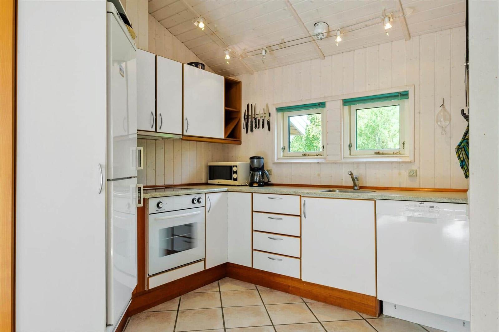 Weiße L-Küche mit Backofen, Geschirrspüler, Kühlschrank und zwei Fenstern.