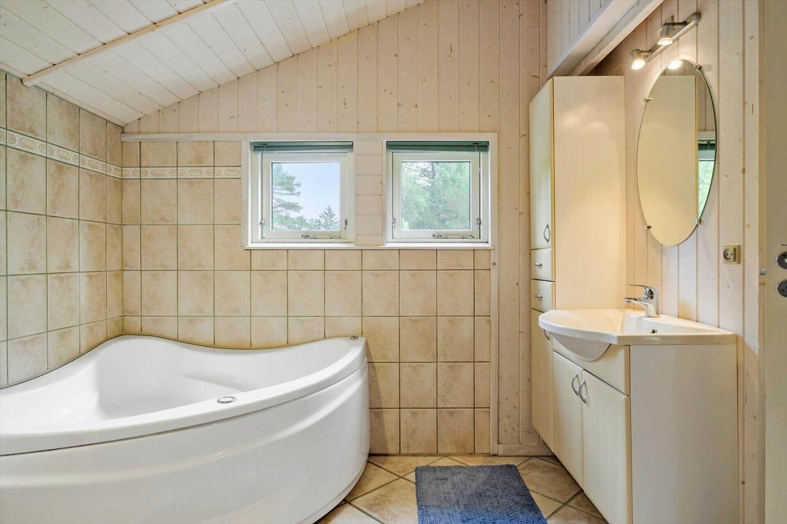 Badezimmer mit Eckbadewanne, Waschtisch, ovalem Spiegel und zwei Fenstern.
