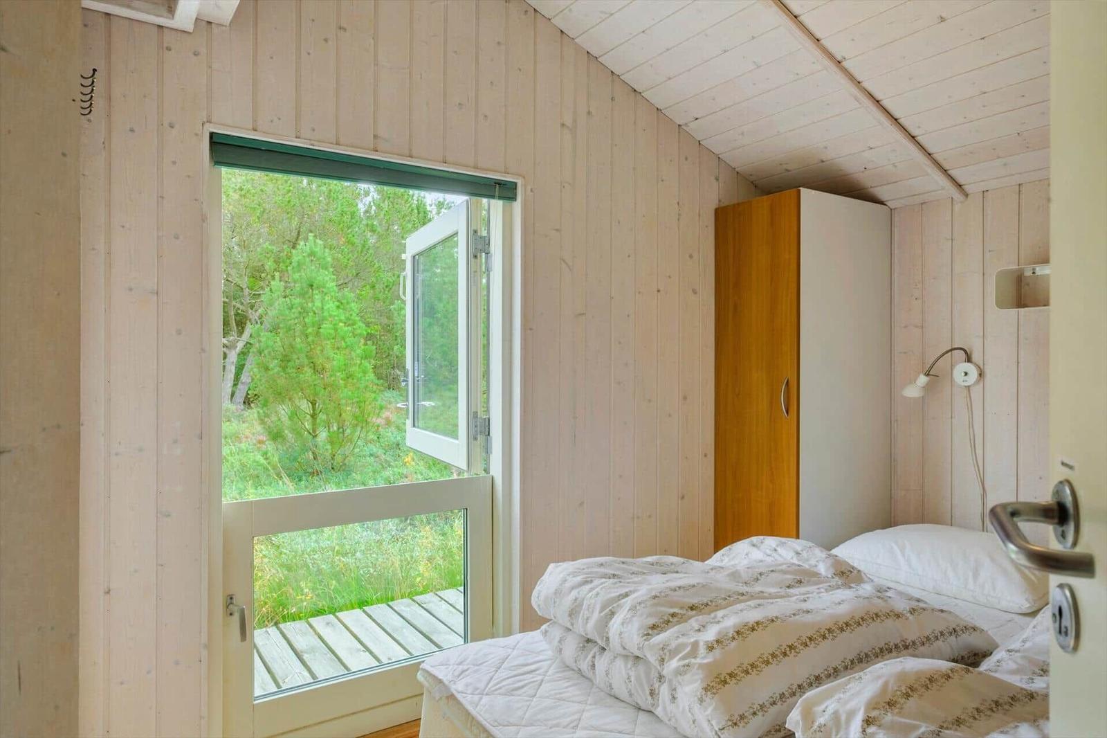 Schlafzimmer mit Holzwänden, Bett, Schrank und offener Terrassentür zu Bäumen.