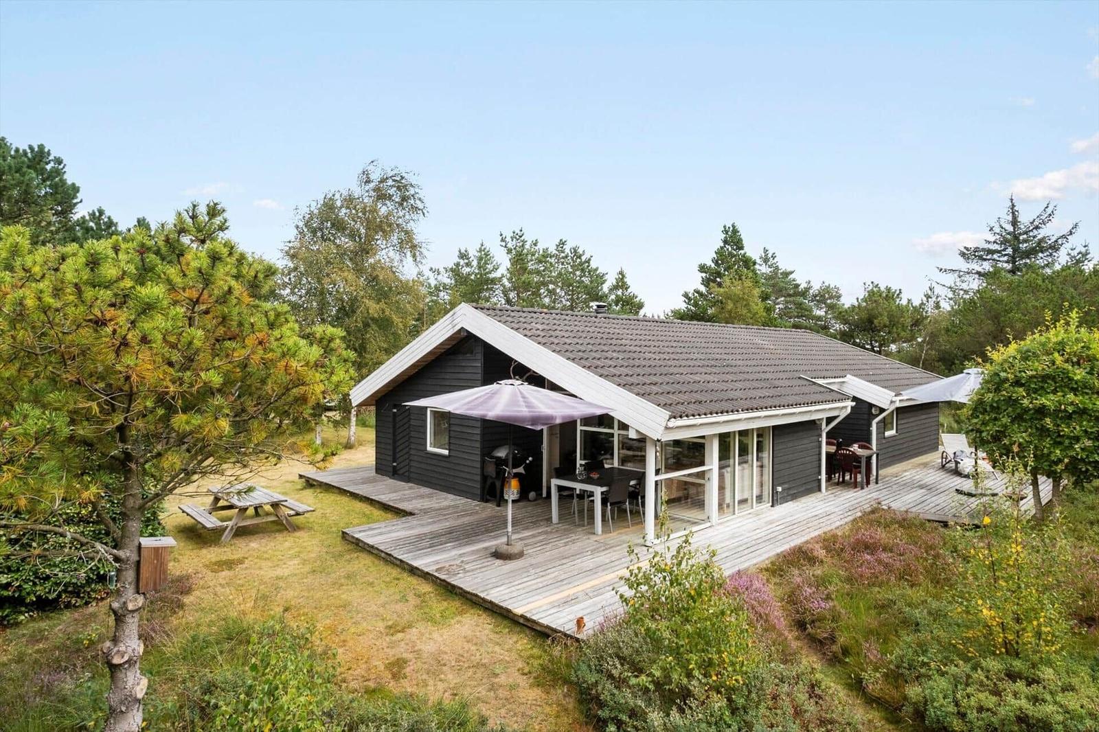 Dunkles Holzhaus mit Terrasse, lila Sonnenschirm, Grill und Picknicktisch.