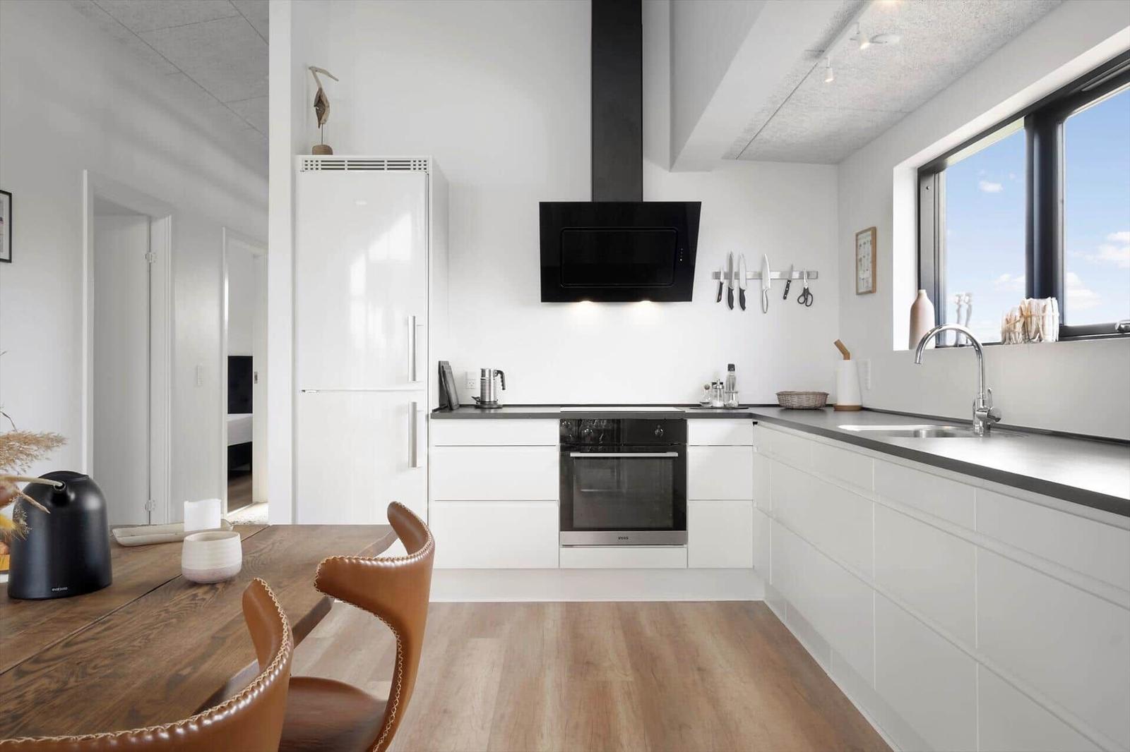 Weiße Küche mit Kühlschrank, Backofen, Dunstabzug und Esstisch mit braunen Lederstühlen.