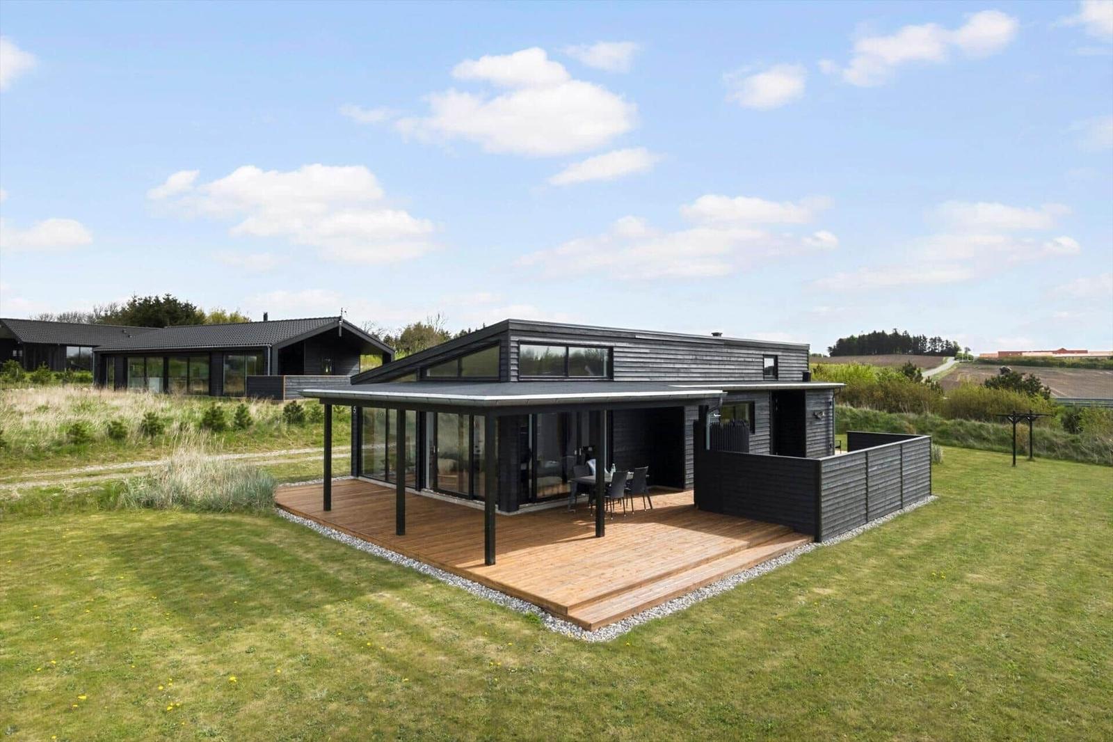 Schwarzes Holzhaus mit großer Holzterrasse, überdachter Sitzgruppe und Rasenfläche.