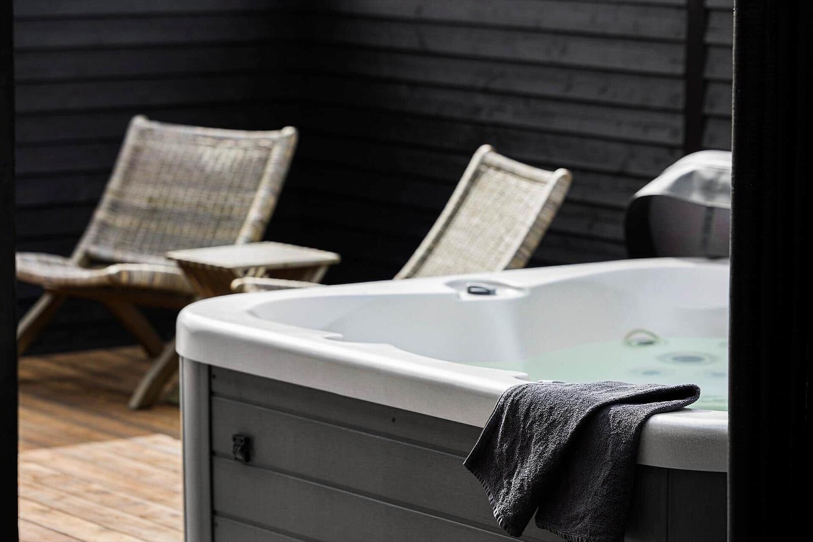 Whirlpool auf Holzterrasse mit Handtuch und zwei Rattansesseln an dunkler Holzwand.