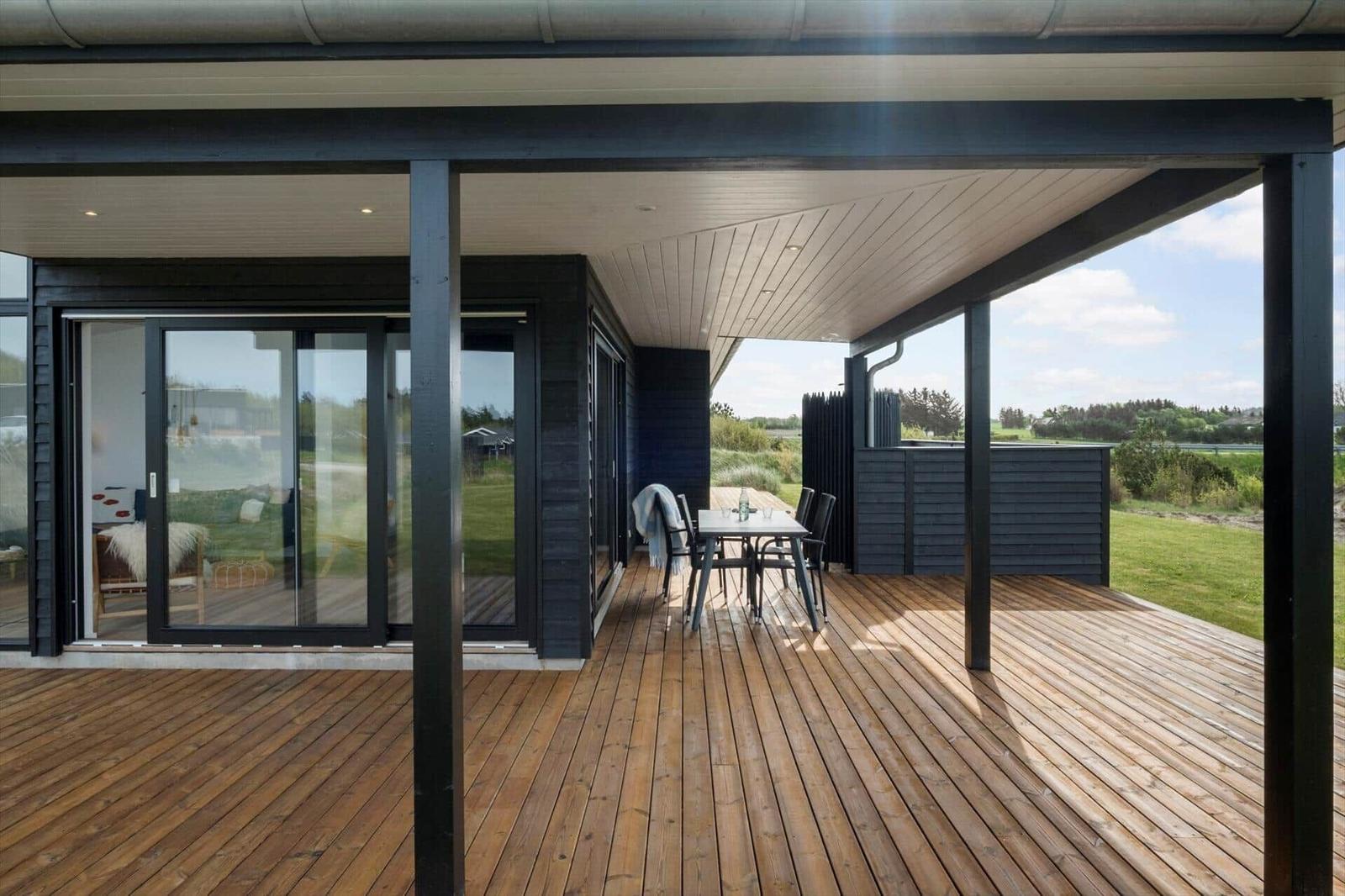 Überdachte Holzterrasse mit Esstisch, Stühlen und Schiebetüren am schwarzen Haus.
