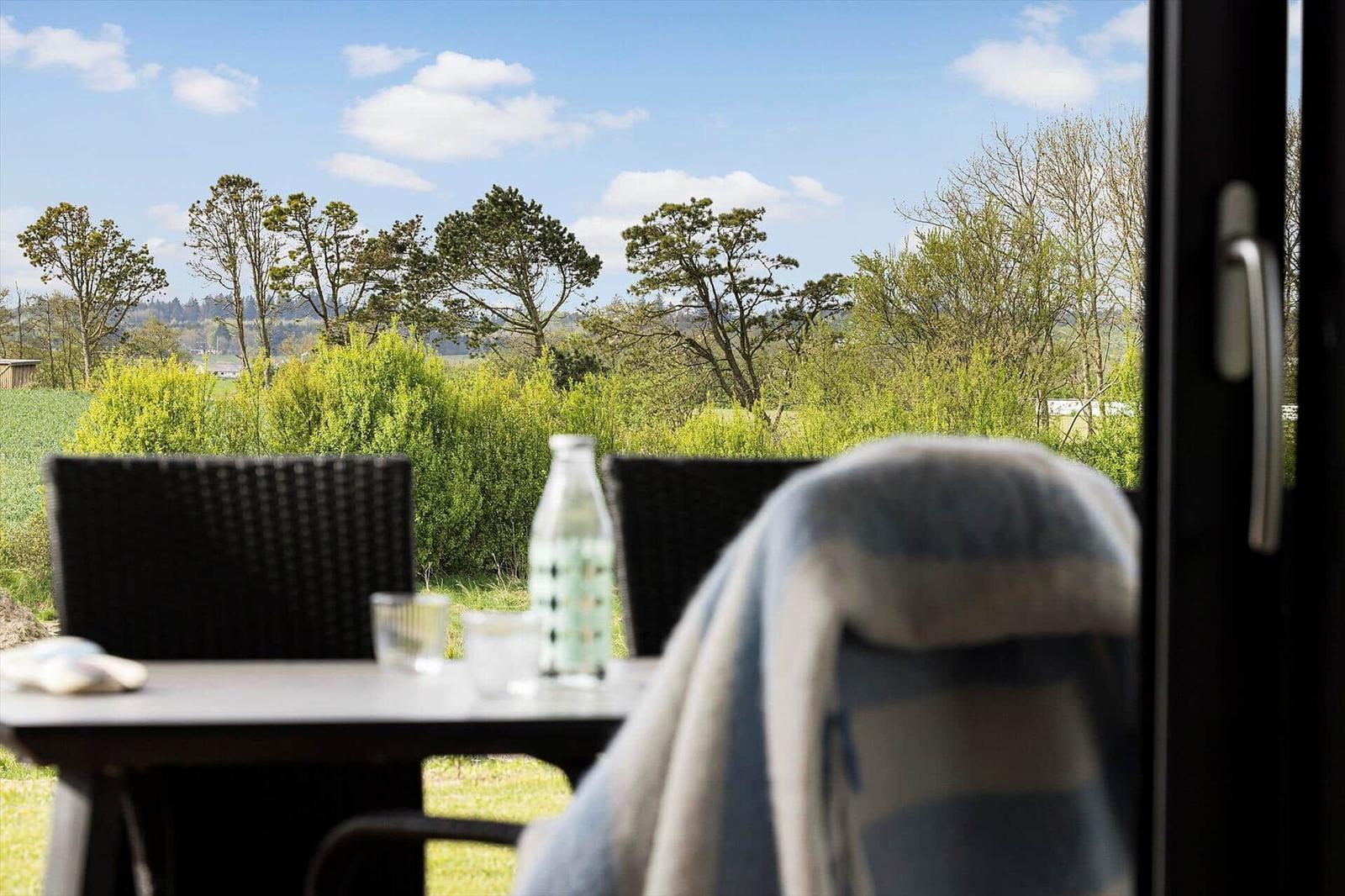 Blick aus dem Haus auf Terrassentisch, Stühle, Hecke, Wiese und Bäume.