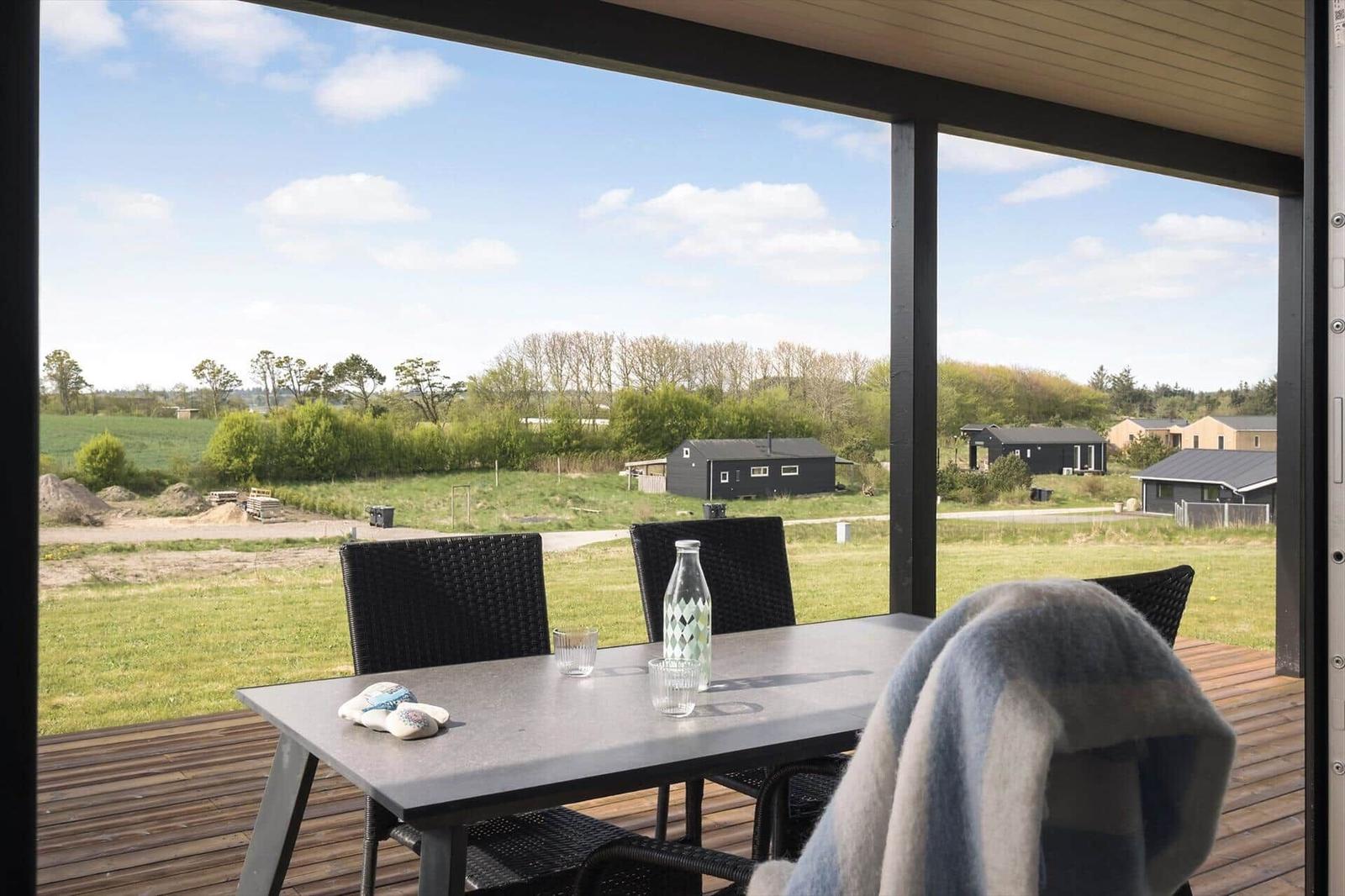 Überdachte Holzterrasse mit Esstisch und Blick auf schwarze Holzhäuser auf der Wiese.