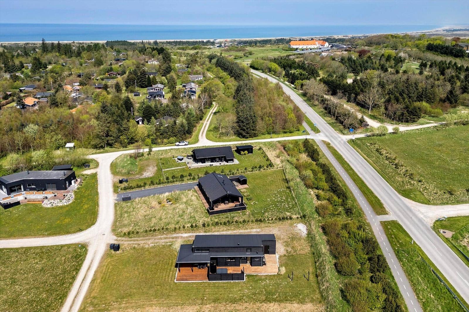 Luftbild schwarzer Ferienhäuser mit Holzterrassen, Straße und Meer am Horizont.