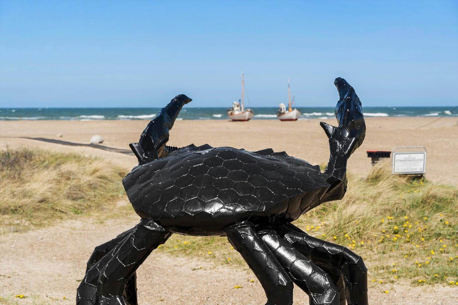 Schwarze Krabbenskulptur im Sand vor Strand, Fischerbooten und Meer.