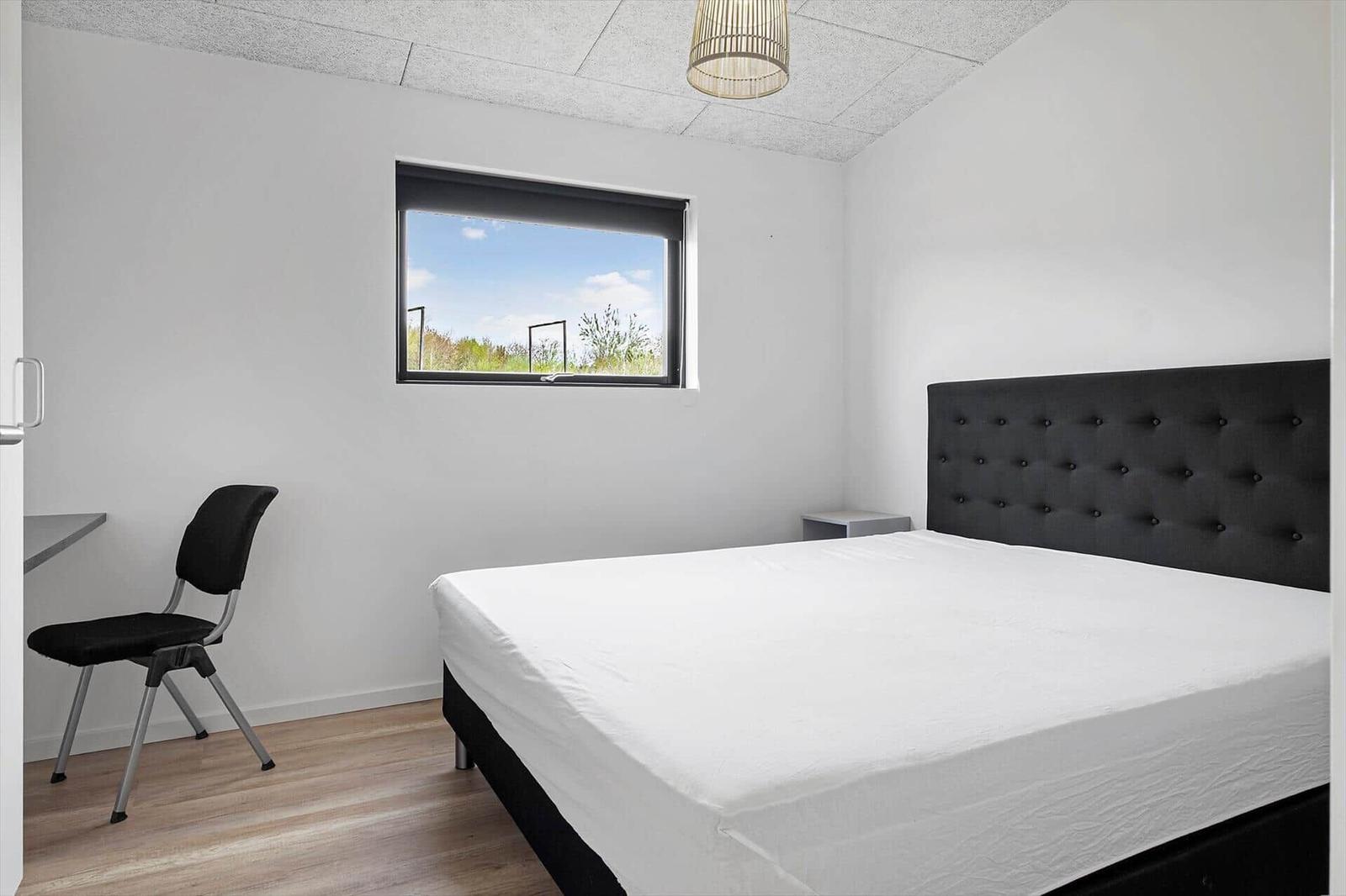 Schlafzimmer mit Doppelbett, dunklem Kopfteil, Schreibtisch und Fenster.