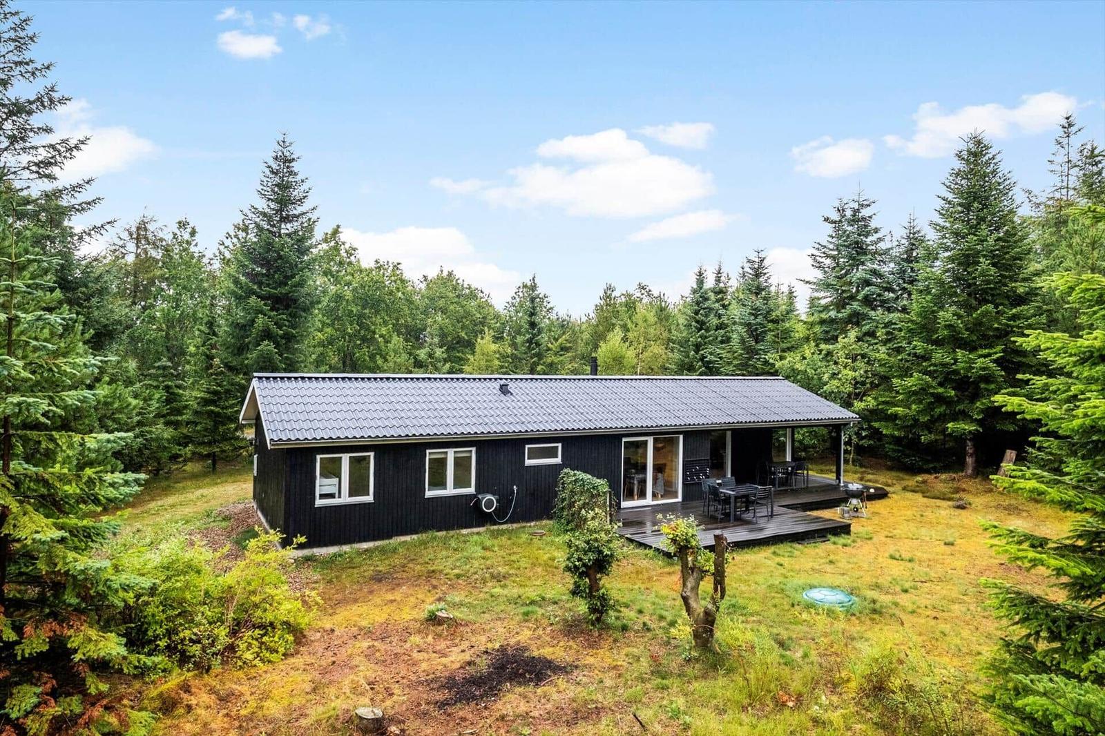Black wooden holiday home with terrace, dining set and grill by the forest.
