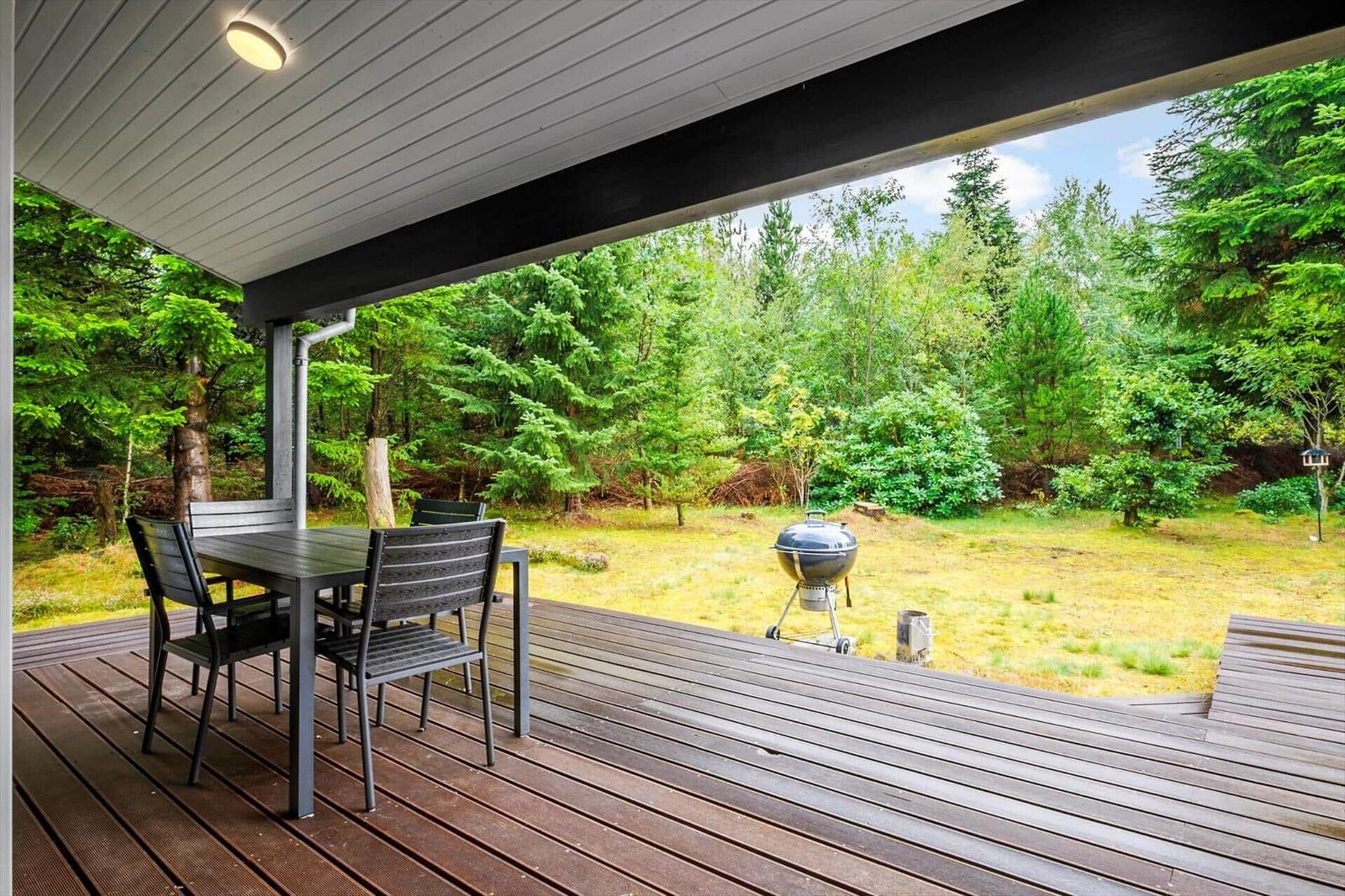 Covered wooden terrace with dining table, lawn view and kettle grill.