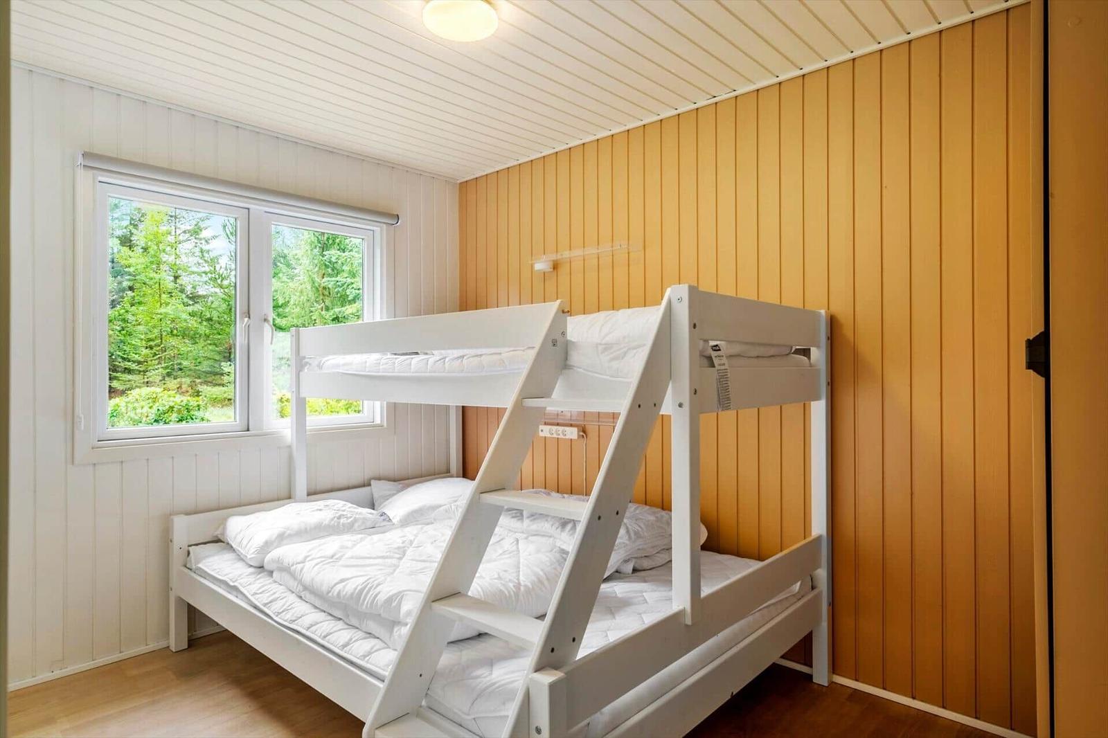 Bedroom with white bunk bed, wood paneling and window view of trees.