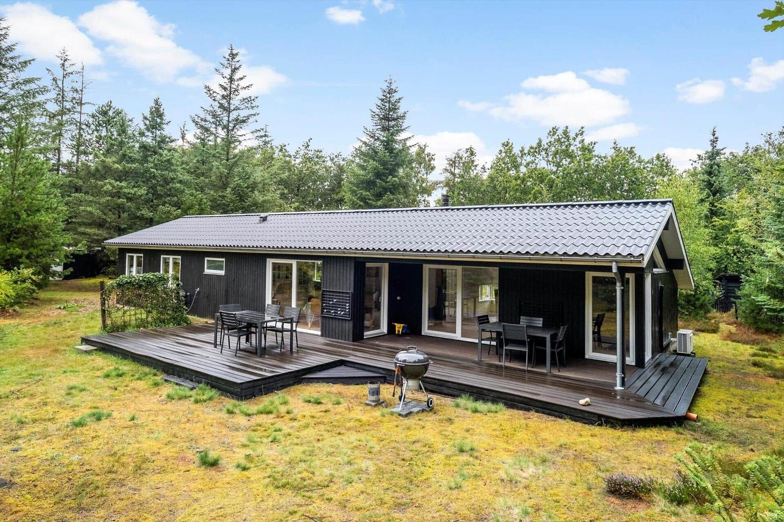Black wooden house with timber deck, garden furniture and grill by the forest.