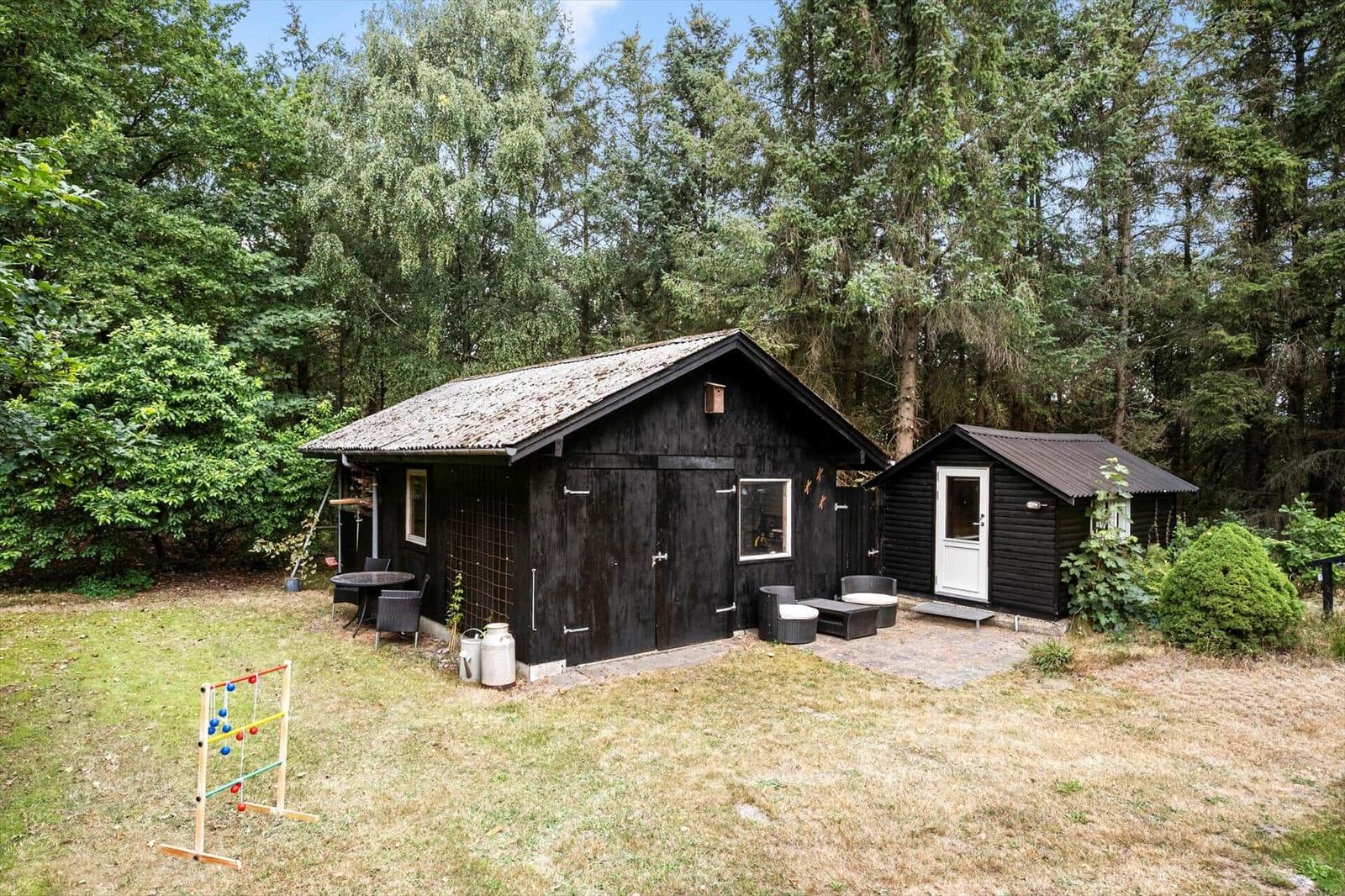 Two black wooden buildings in the forest with seating, lawn and a tossing game in front.