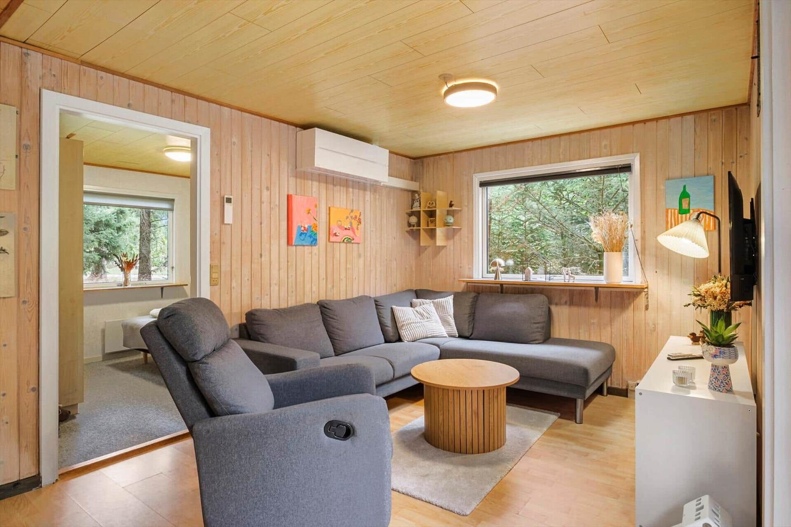 Living room with grey corner sofa, round coffee table, armchair and TV facing a forest window.