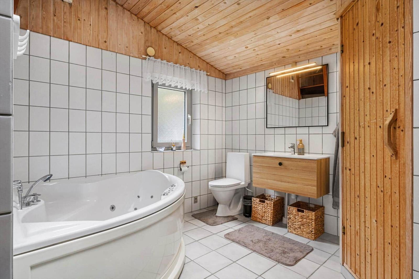 Bathroom with corner whirlpool tub, toilet, wooden vanity, mirror and white tiled walls.