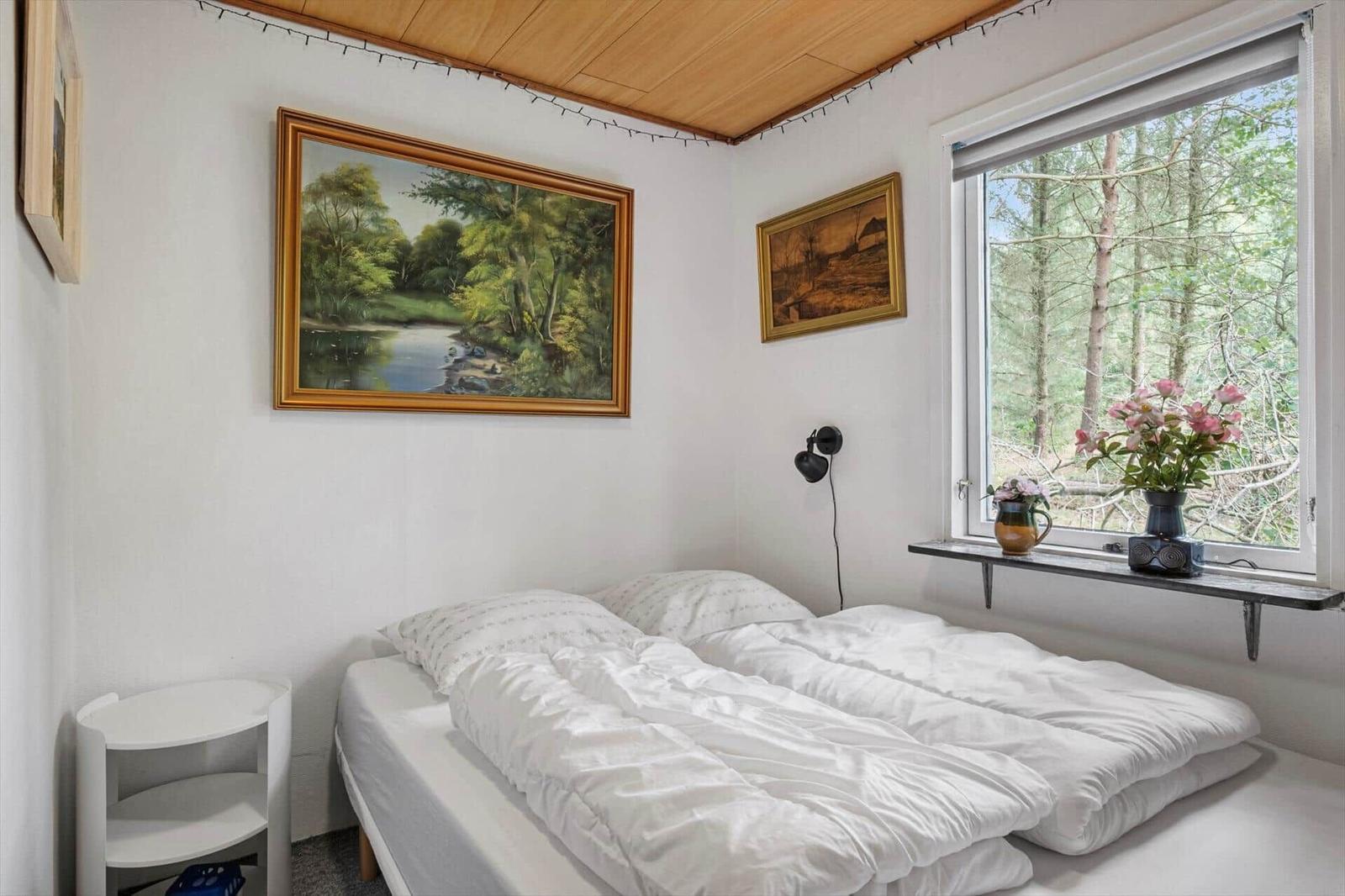 Bedroom with double bed, white bedding, landscape paintings and window with a forest view.