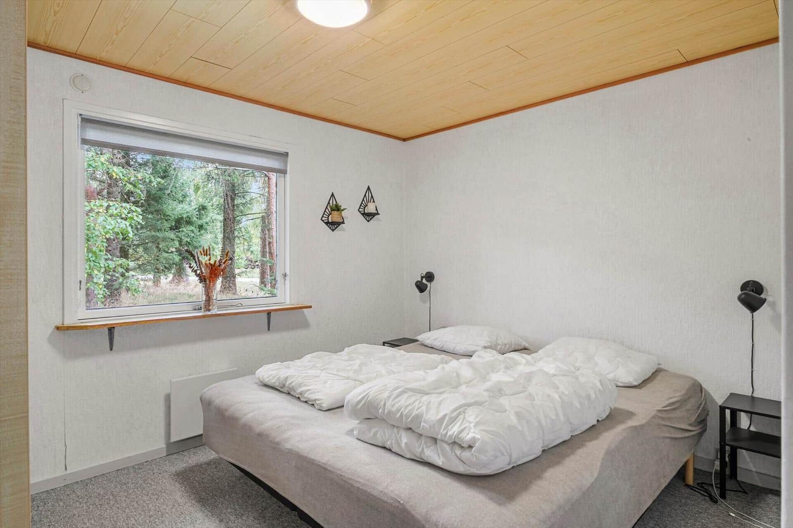 Bedroom with double bed, reading lamps, carpet and a large window with forest view.