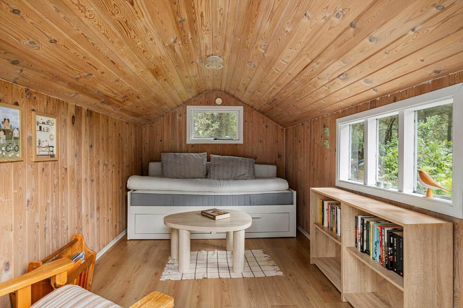Wood-paneled room with daybed, round coffee table, bookshelf and forest windows.