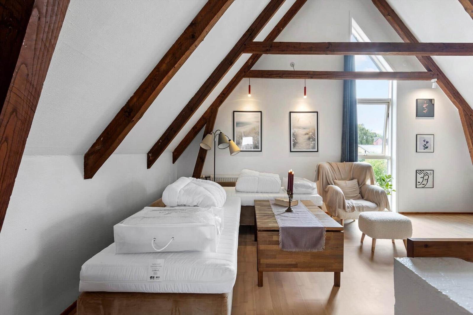 Attic room with two single beds, wooden table, armchair, stool and wooden beams.