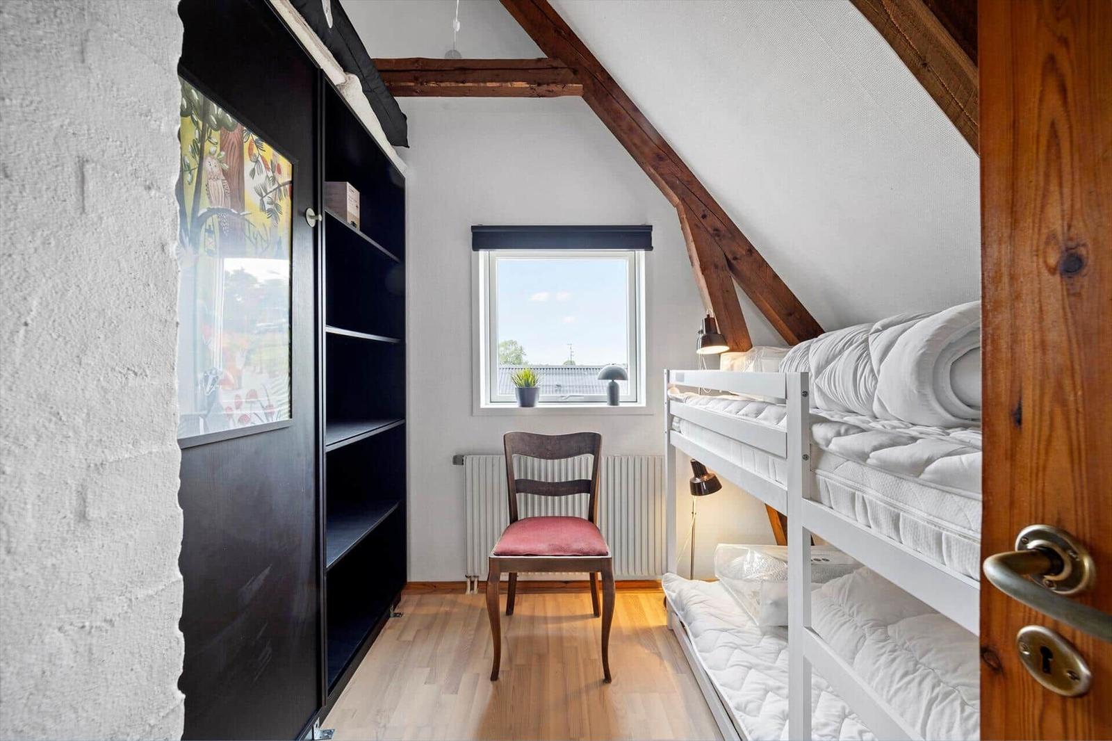 Attic room with white bunk bed, wooden chair, black shelf and small window.