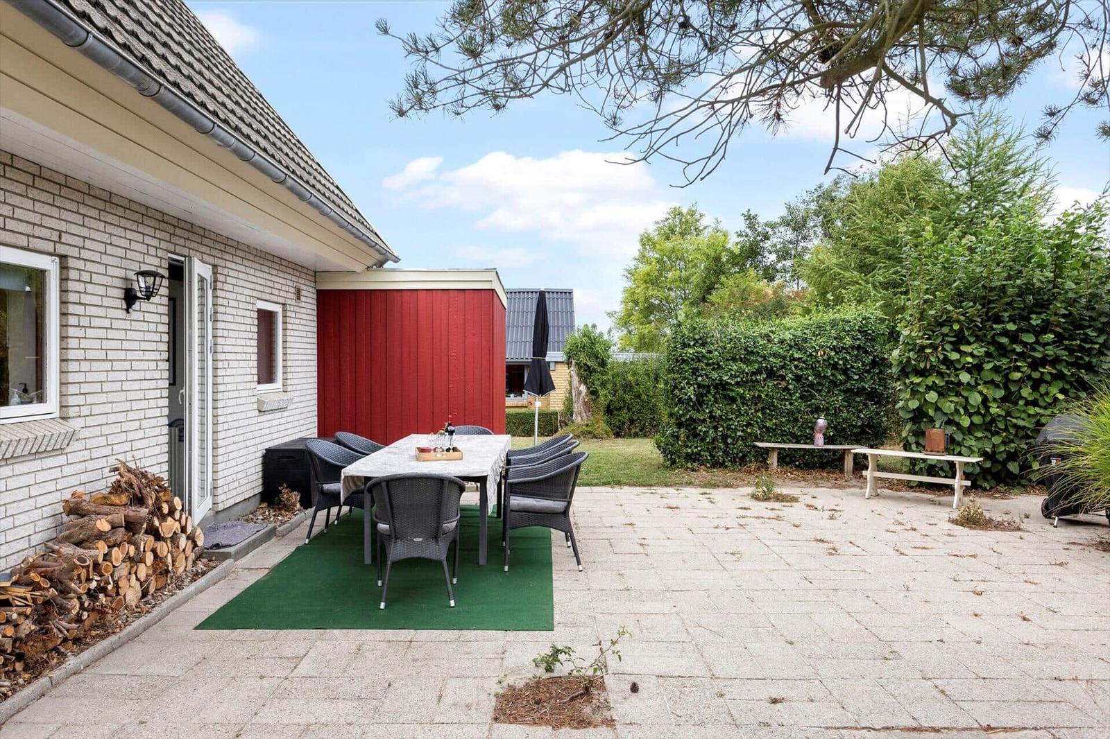 Terrace with dining table, wicker chairs, firewood stack and hedge by the house.