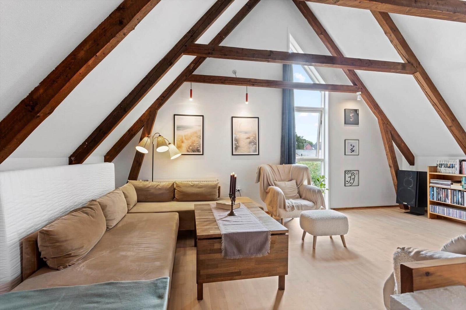 Attic living room with brown corner sofa, wooden table, armchair and exposed beams.