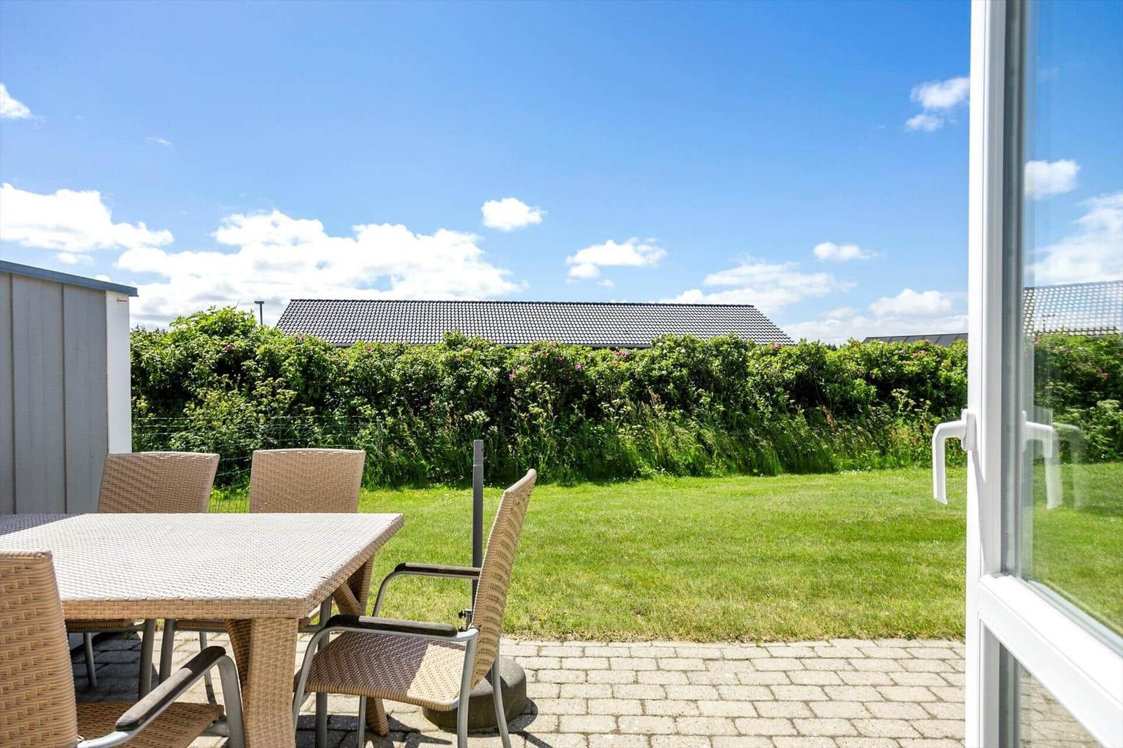 Terrasse mit Gartenmöbeln, Pflaster, Rasen und Hecke unter blauem Himmel.
