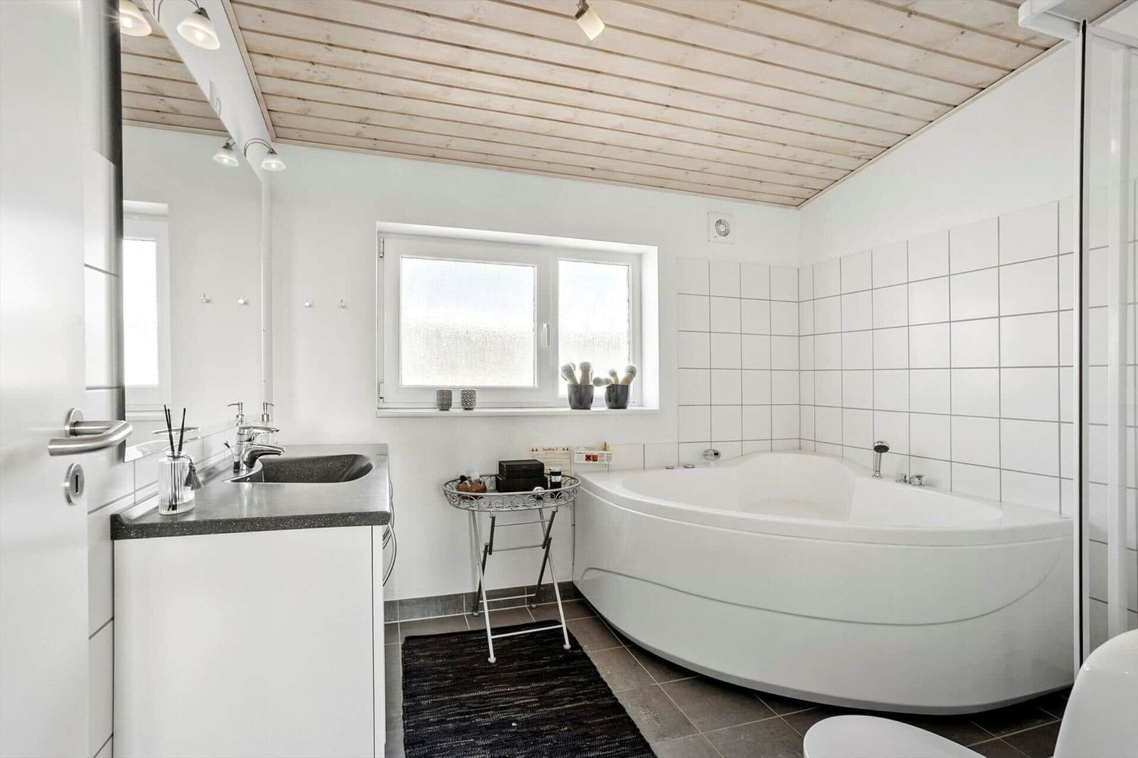 Badezimmer mit Eckbadewanne, Waschtisch, Spiegel, Fenster und Holzdecke.