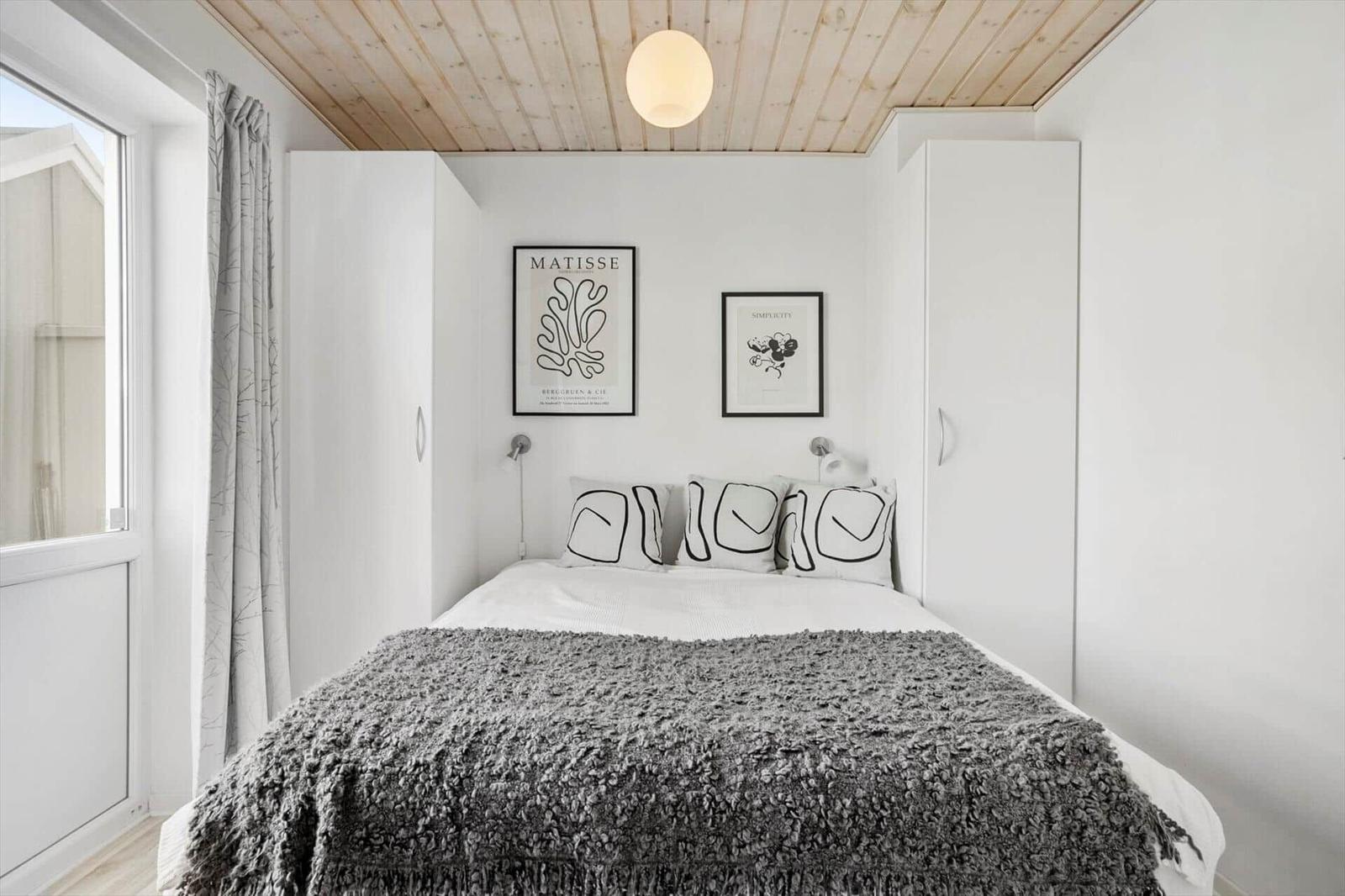 Schlafzimmer mit Doppelbett zwischen weißen Schränken, grauer Tagesdecke und Holzdecke.