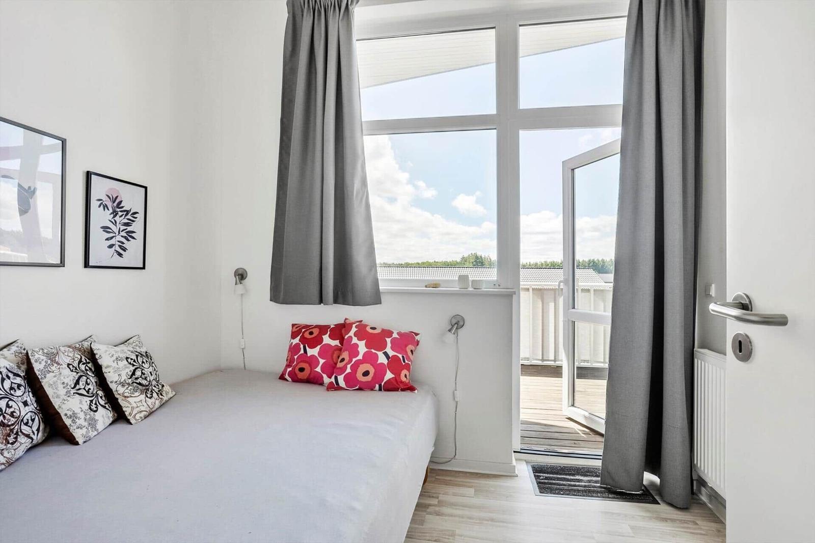 Schlafzimmer mit Bett, Blumenkissen, offener Balkontür und hellem Holzboden.
