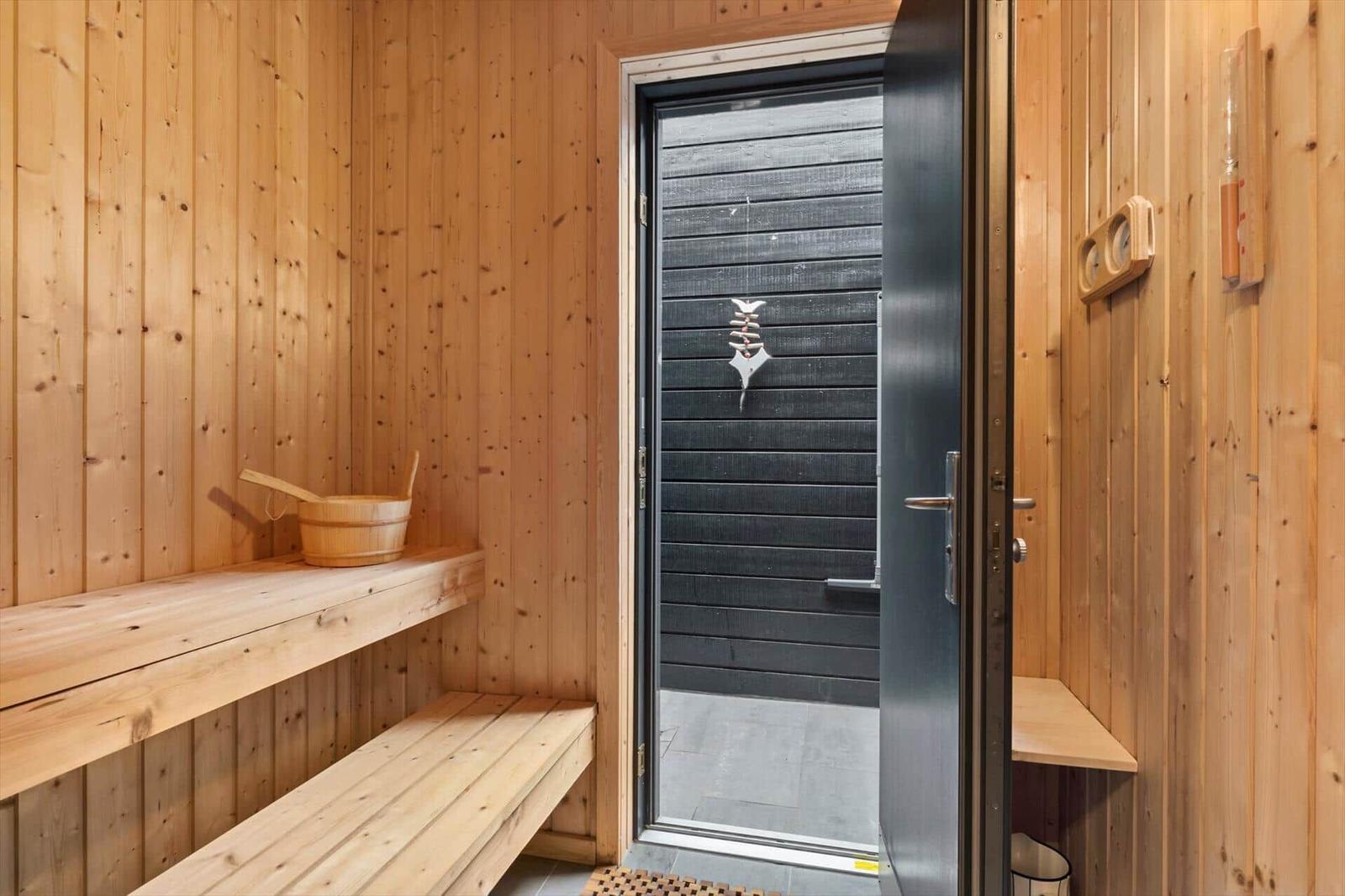 Wooden sauna with two tiers of benches, bucket and open door to the patio.