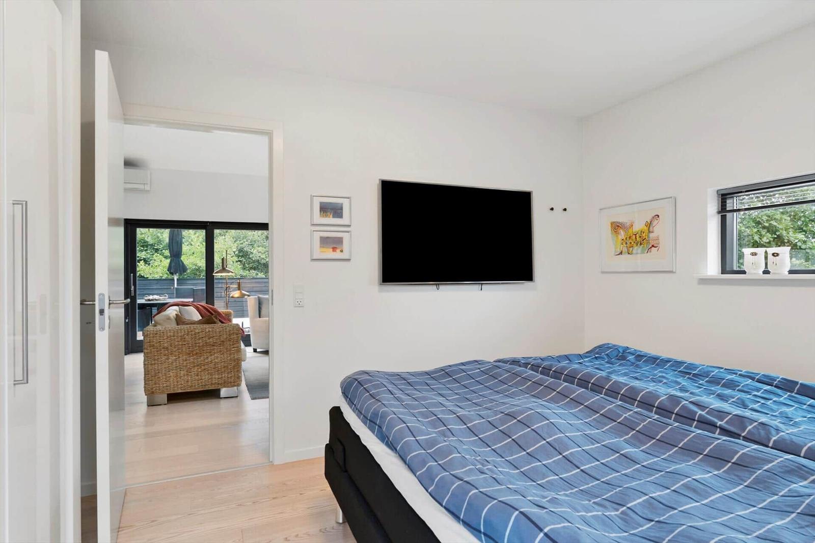 Bedroom with checkered bedding, wall-mounted TV, pictures and view into the living room.