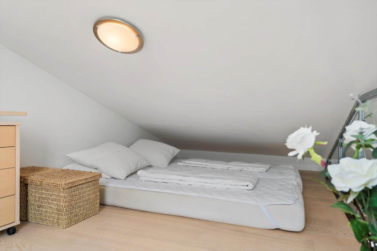 Sleeping loft under a sloped ceiling with mattress, white bedding and wicker basket.