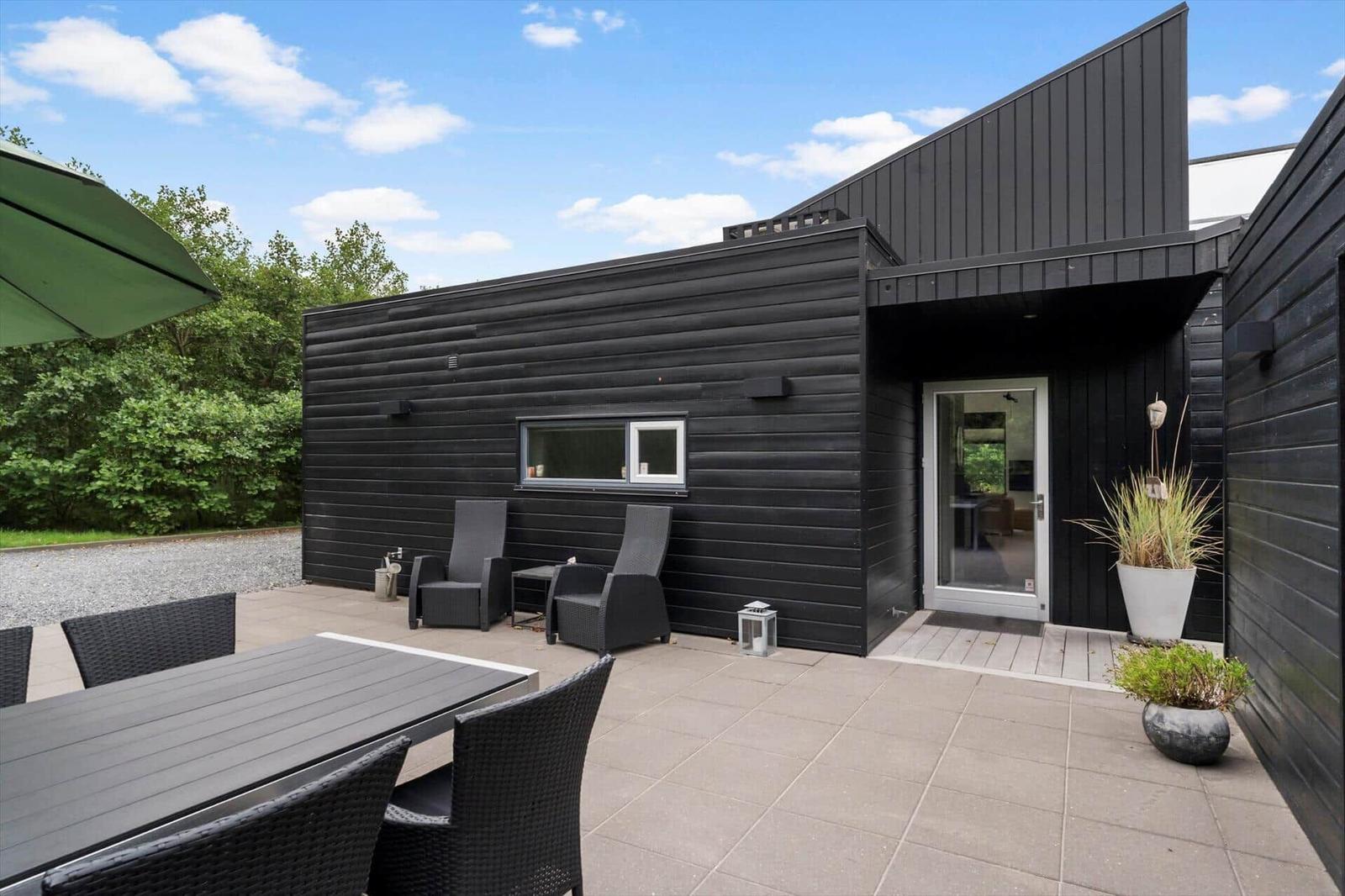 Stone terrace by a black wooden house with lounge chairs, dining table and white door.