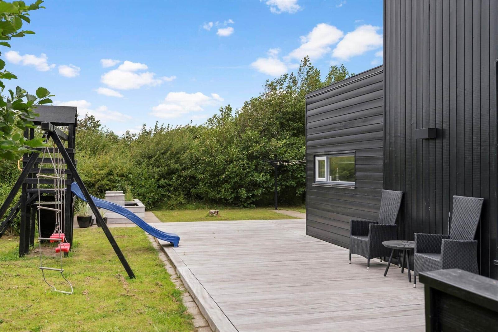 Wooden terrace with black chairs, lawn and climbing frame with a blue slide.