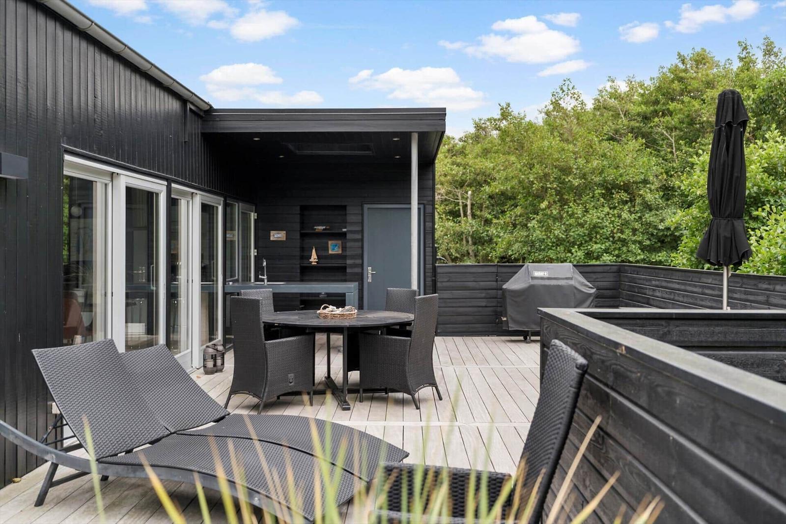 Wooden terrace with sun loungers, round dining table, grill and black wood facade.