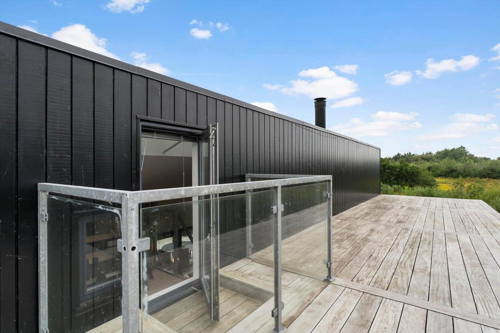 Wooden terrace with glass railing, black wood facade, chimney and open door.