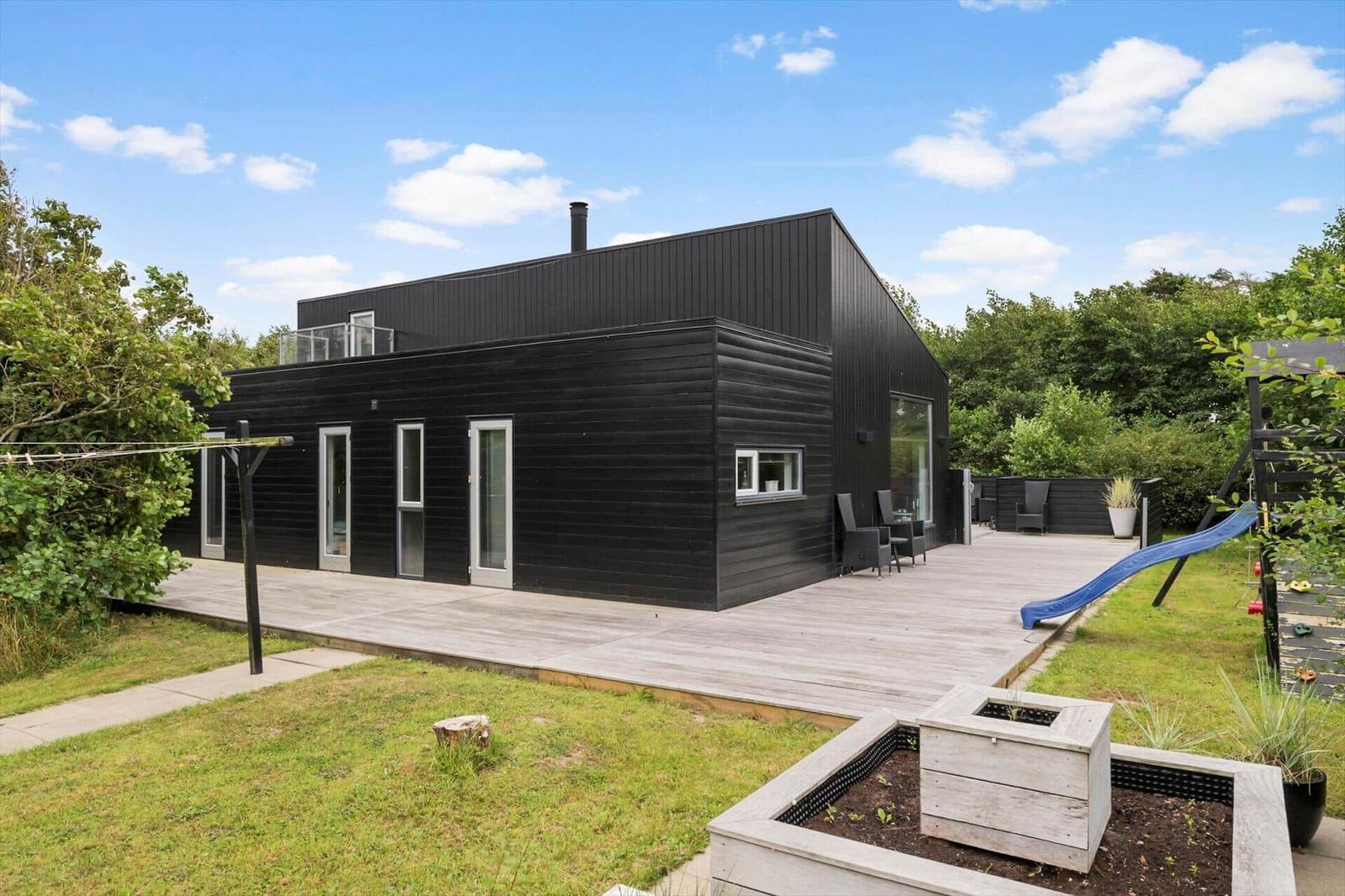 Black wooden house with terrace, garden furniture, lawn and play set with a slide.