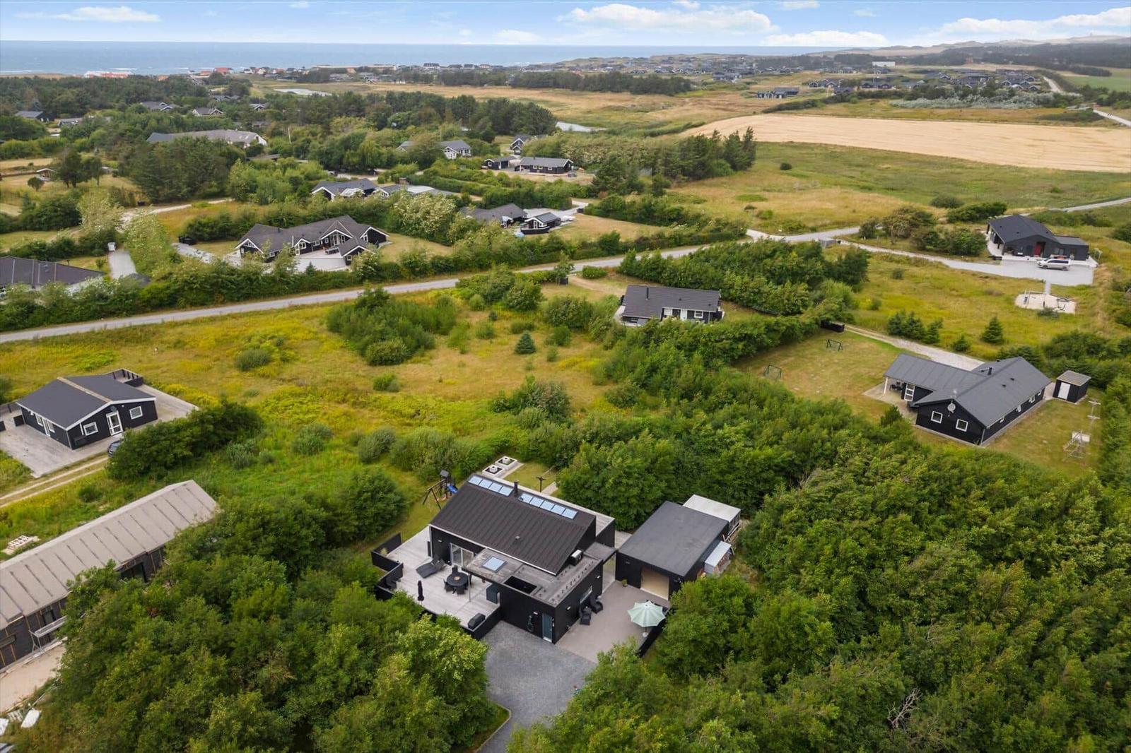 Aerial view of a black house with wooden terrace, yellow field and sea on the horizon.
